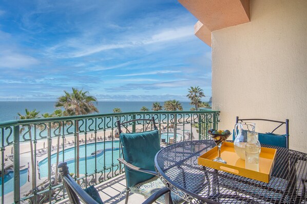 Un balcon avec vue sur une piscine et une plage, une table avec un plateau, et une chaise.