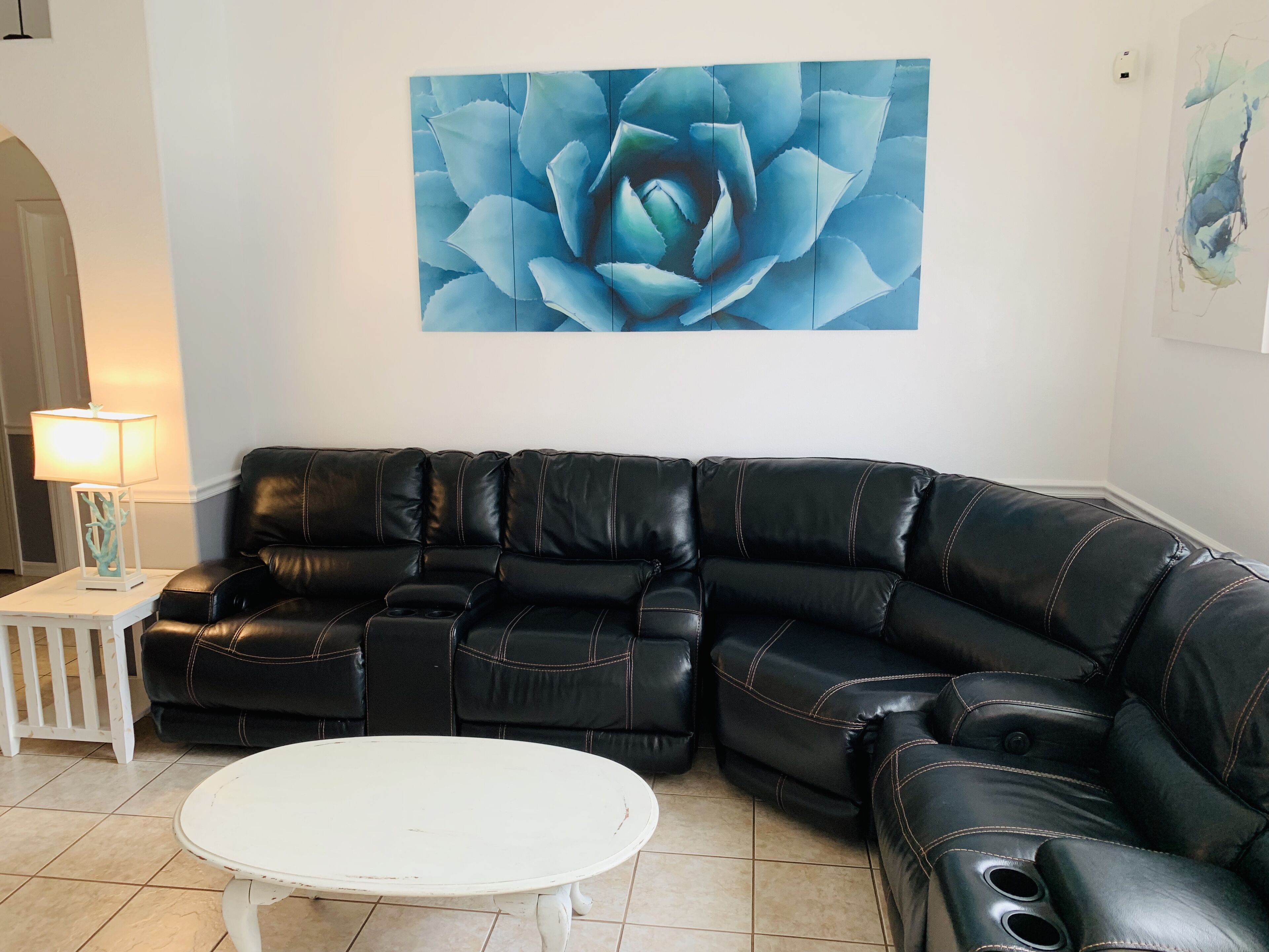 En stue med en sort lædersofa, et hvidt sofabord og et stort blå blomstermaleri på væggen.