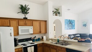 A modern kitchen with wooden cabinets, a white refrigerator, a microwave, a stove, and a dishwasher. There are plants on the countertops and a black leather couch in the adjacent living area. - PROMO 10% WEEKLY/15% MONTHLY DISCOUNT
Your Magical Oasis Awaits, Disney Is near. (Kissimmee)