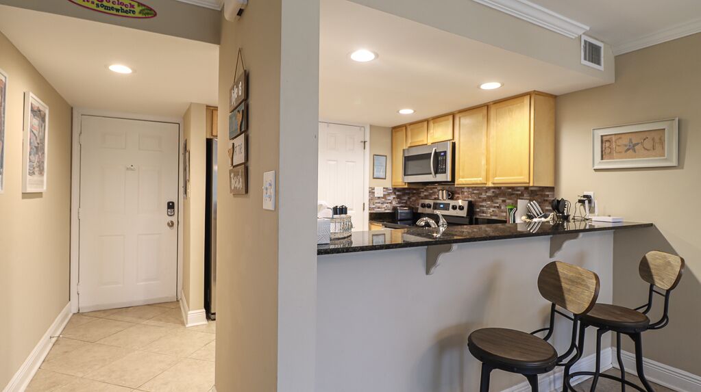 A kitchen with wooden cabinets, a microwave, and a coffee maker. There are two bar stools and a refrigerator.