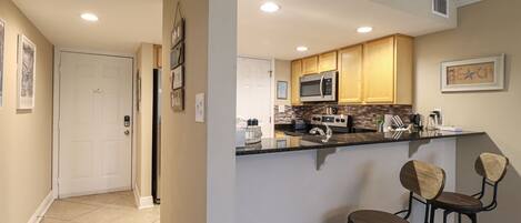 A kitchen with wooden cabinets, a microwave, and a coffee maker. There are two bar stools and a refrigerator.