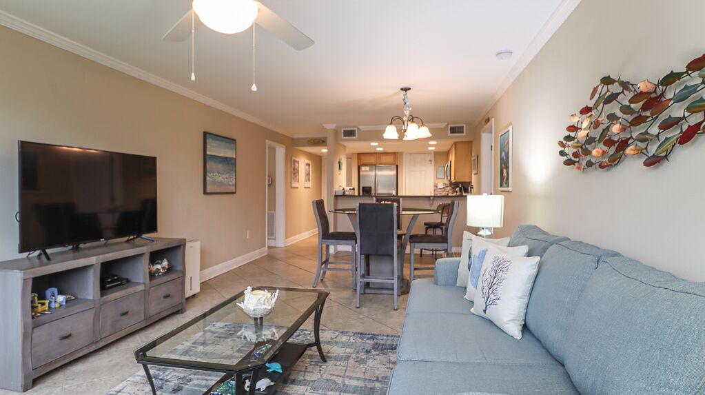 A living room with a TV, sofa, coffee table, and a dining area with chairs and a chandelier.