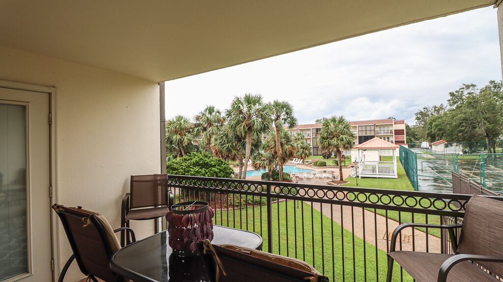 A balcony with a table and chairs, overlooking a pool area with palm trees and a building in the background.