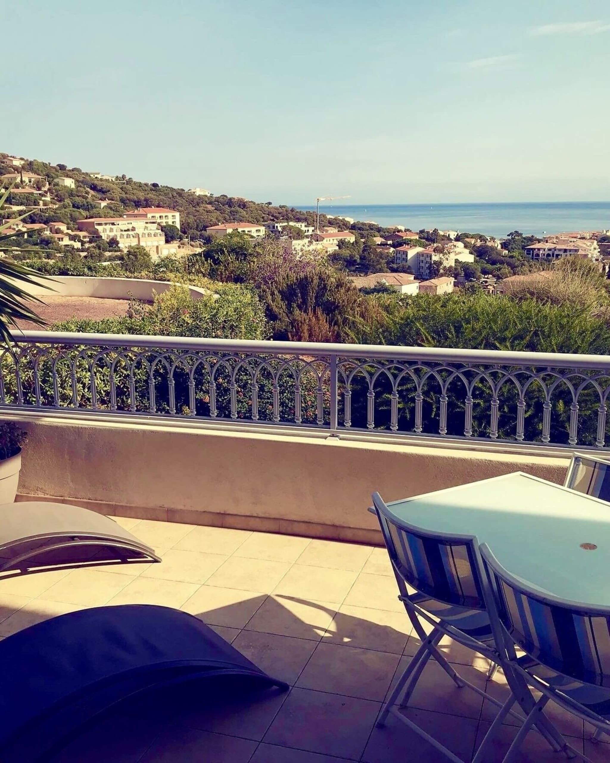 A terrace with a table and chairs, overlooking a coastal town with buildings and a clear sky.