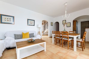 A modern living room with a sofa, coffee table, dining table, and chairs. There is a staircase leading to an upper floor.
