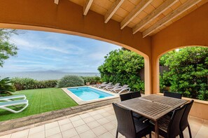 A covered patio with a pool, lounge chairs, and a view of the sea.