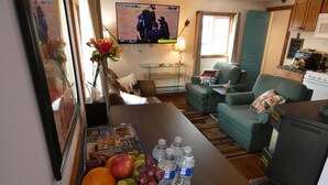 Ein Wohnzimmer mit Fernseher, einem Esstisch mit Obst und Wasserflaschen, einer Couch und einer Küchenzeile.