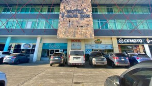 Un hôtel à la façade moderne, un parking où sont garées plusieurs voitures, et différentes boutiques, dont un salon de manucure et un restaurant.