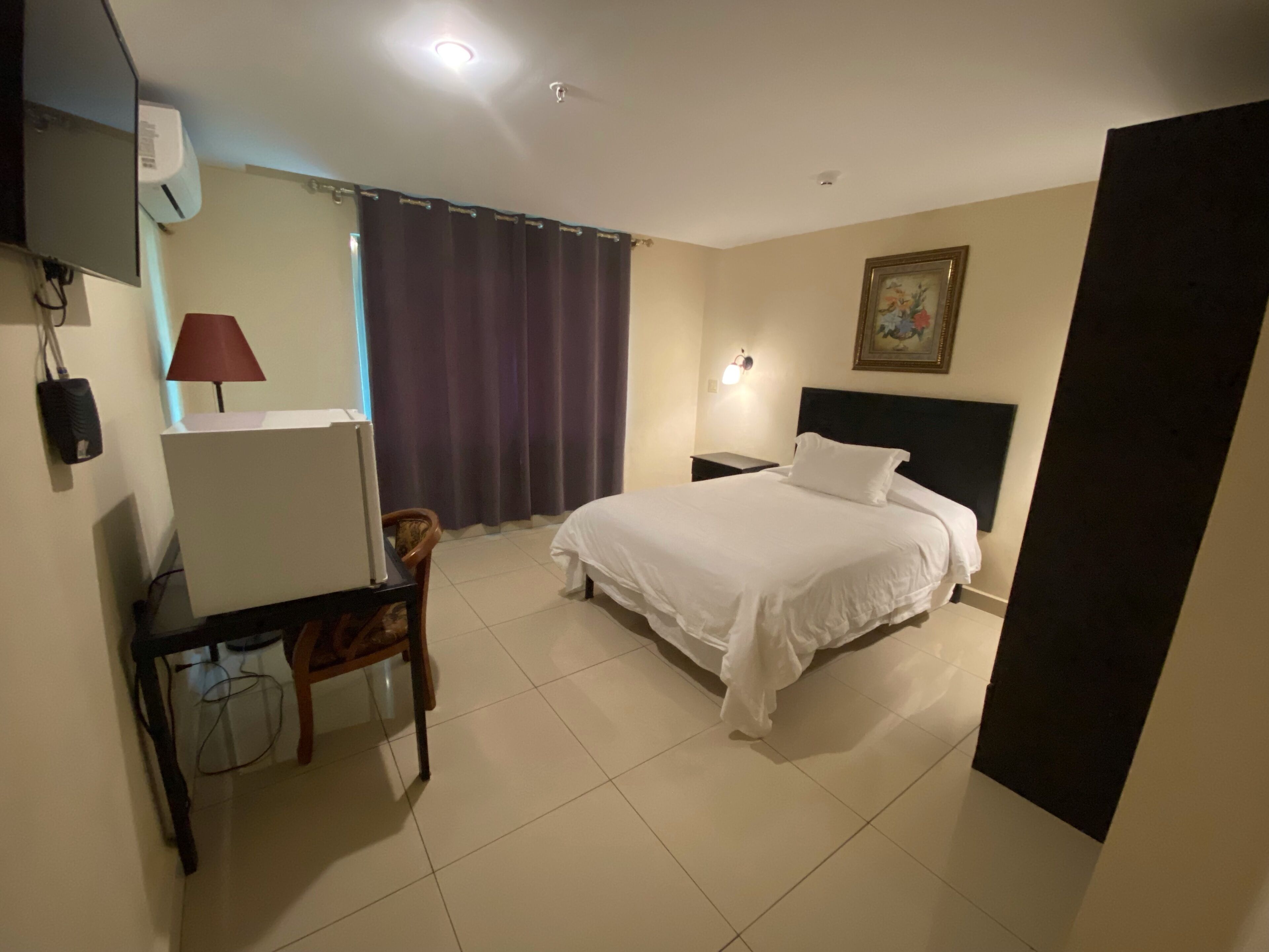 Habitación de hotel con cama, escritorio con silla, mesita de noche, lámpara y un cuadro en la pared.