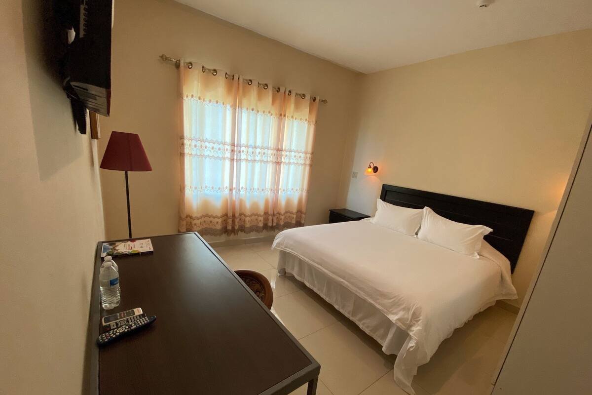 A hotel room with a bed, a desk with a remote control, a lamp, and a window with curtains.