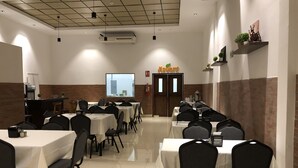 Une salle de conférence avec plusieurs tables et chaises, un écran de projection et un coin cuisine à l’arrière-plan.