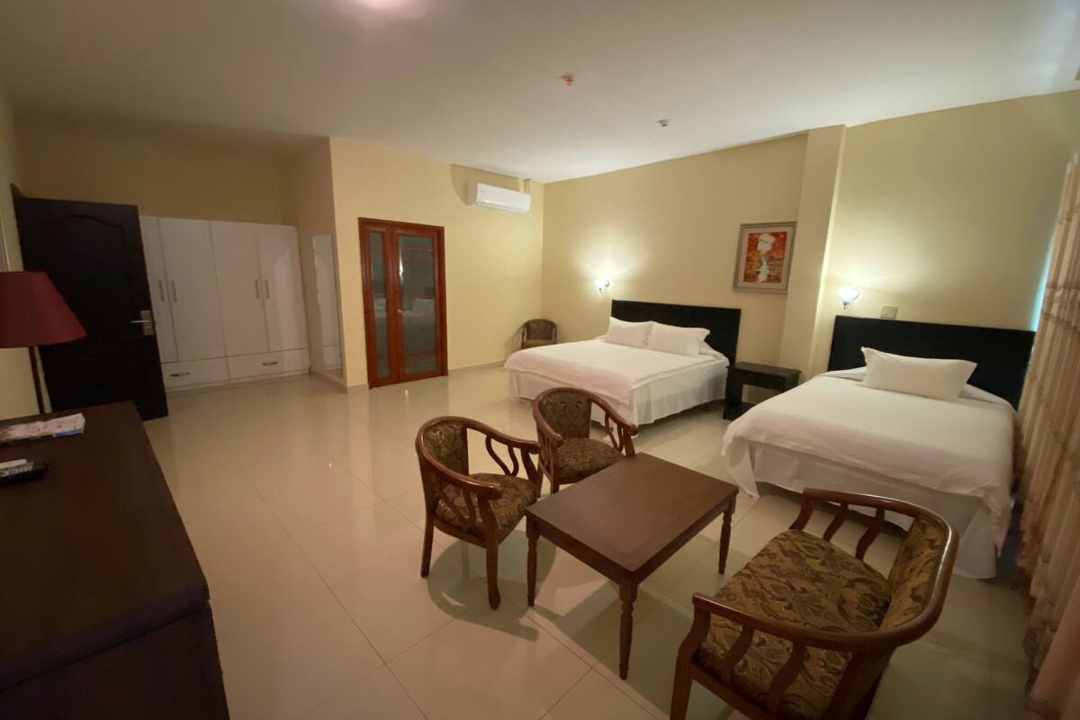 A hotel room with two beds, a desk, and two chairs. There is a wardrobe, a door, and a wall-mounted air conditioner.