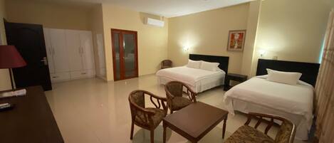 A hotel room with two beds, a desk, and two chairs. There is a wardrobe, a door, and a wall-mounted air conditioner.
