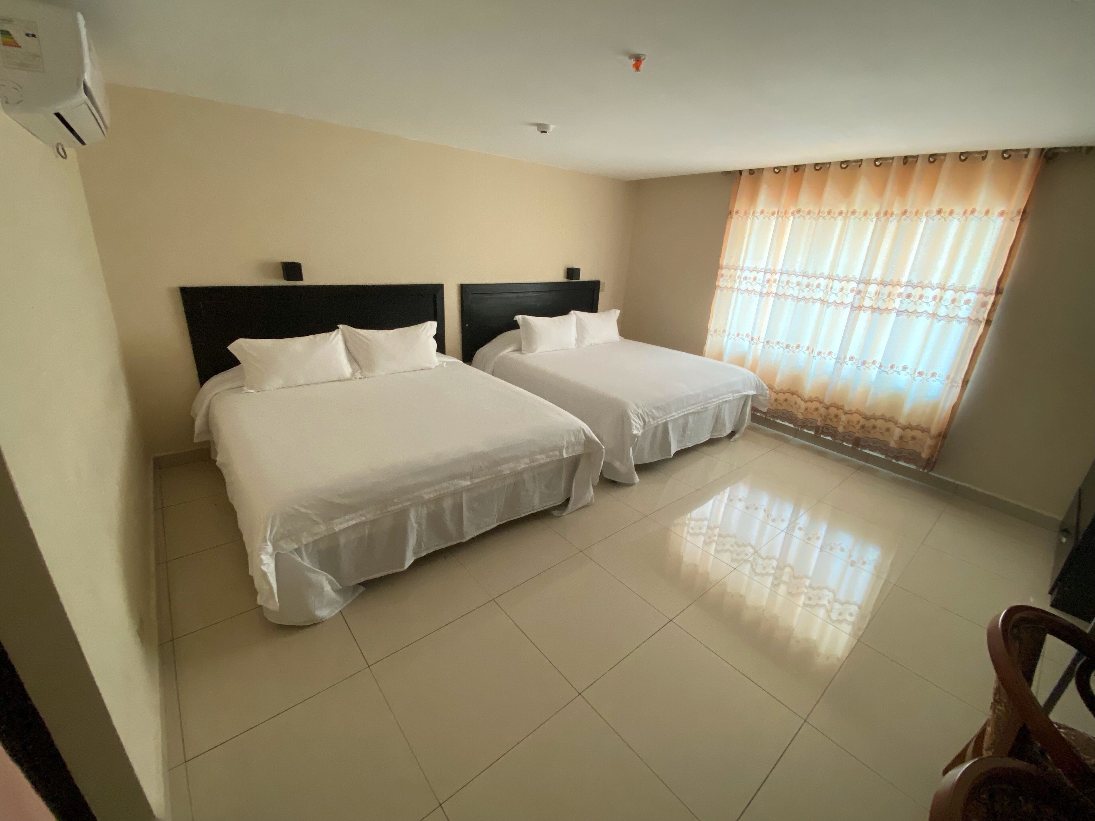 A hotel room with two beds, a tiled floor, a ceiling fan, and a window with curtains.