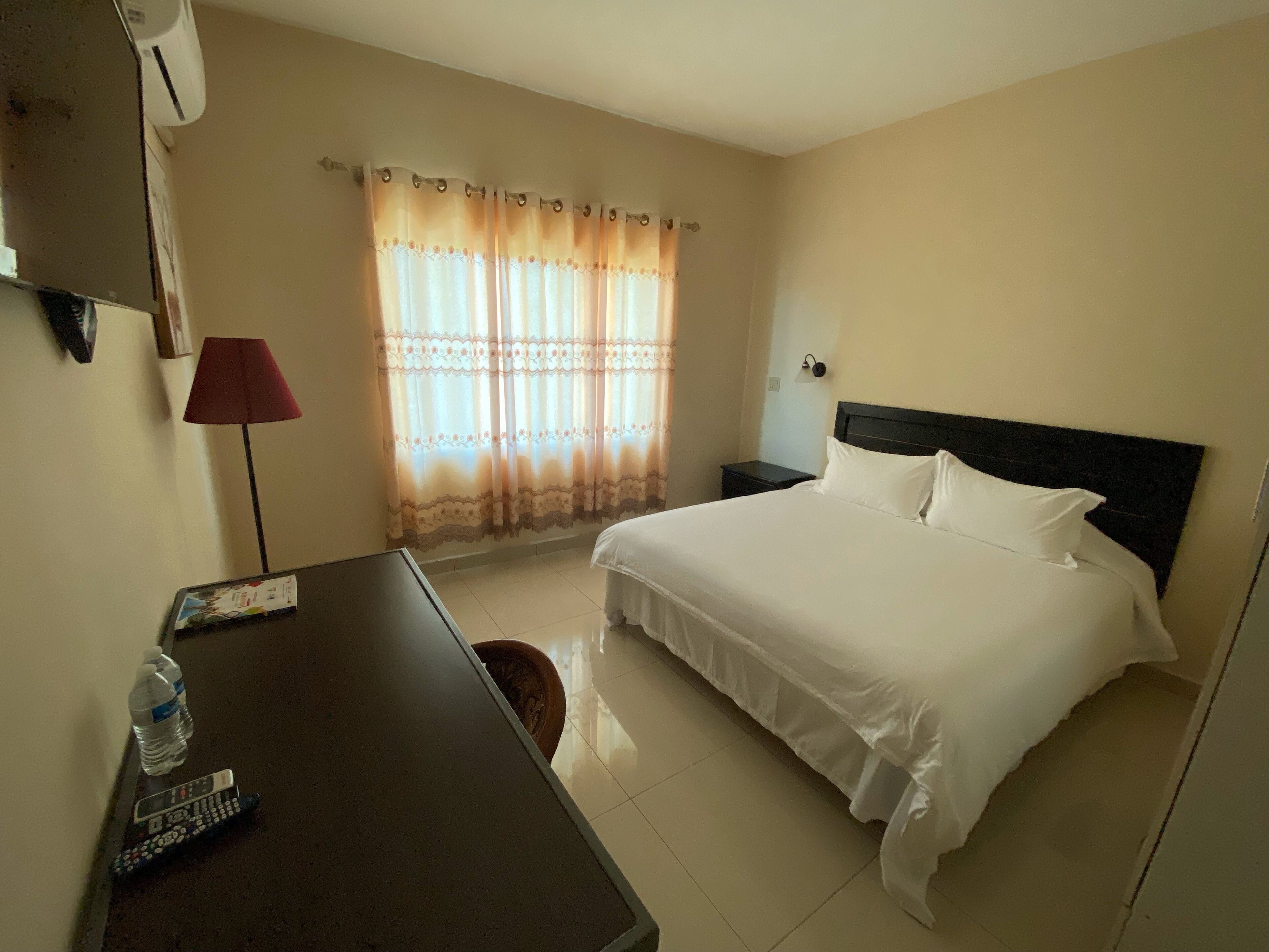 Habitación de hotel con cama, escritorio, lámpara, televisor y ventana con cortinas.