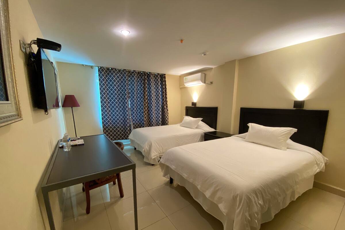 A hotel room with two beds, a desk, a chair, a lamp, and a window with curtains.