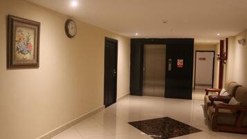 A hotel lobby with a tiled floor, a clock, a framed floral artwork, an elevator, and a seating area with chairs.