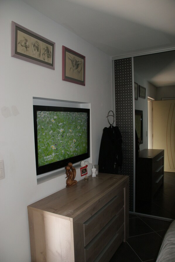 Un dormitorio con televisor montado en la pared, cómoda de madera, perchero y armario corredero.