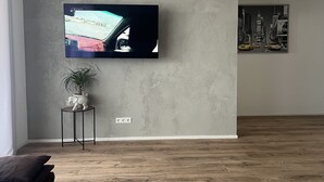 Moderni olohuone, jossa on seinälle asennettu litteänäyttöinen televisio, seinän taustalla on kuvioitu harmaa pinta. Huoneessa on pieni sivupöytä, jonka päällä on kasvi ja koriste-esineitä, sekä puulattia.