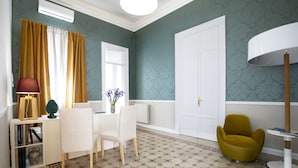 Una habitación con piso estampado, un escritorio blanco con una lámpara, una silla, una mesita con un florero y un sillón verde.