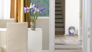 Una habitación con una silla blanca, una pequeña mesa con un jarrón de flores, un papel tapiz decorativo y una escalera al fondo.