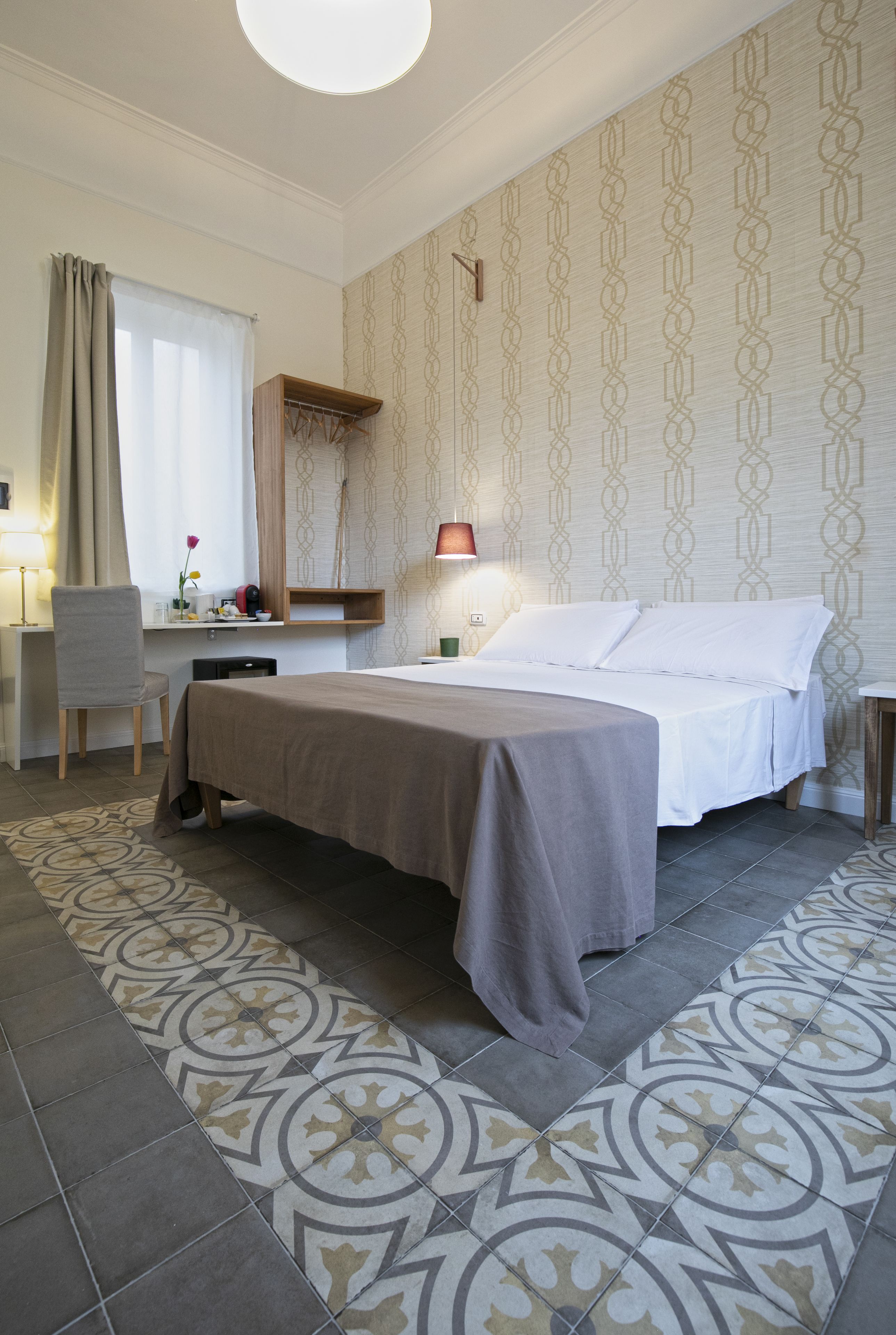 A hotel room with a bed, a desk, a chair, a lamp, and patterned wallpaper.