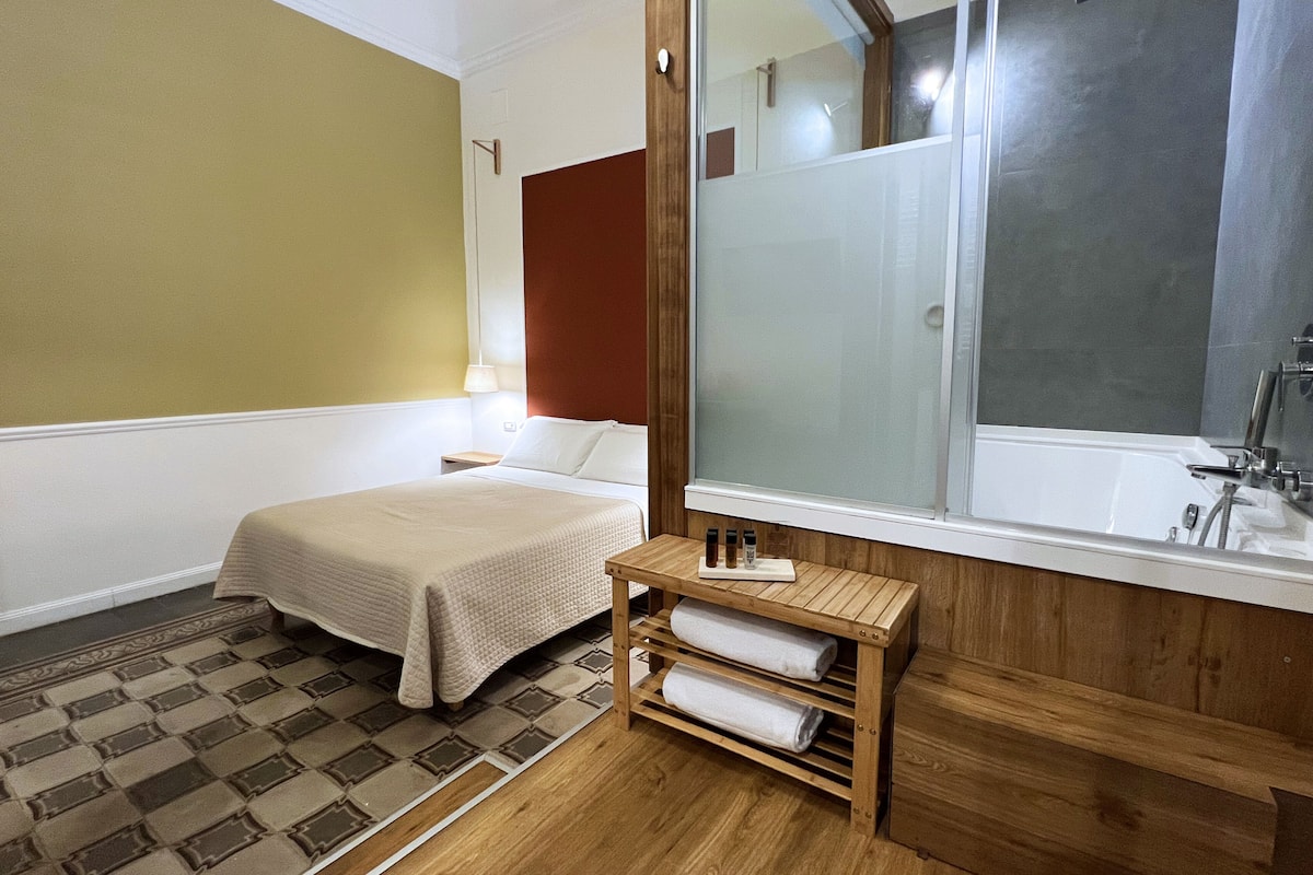 Habitación de hotel con cama, una pequeña mesa de madera con toallas, mesita de noche, espejo y baño con bañera y ducha.