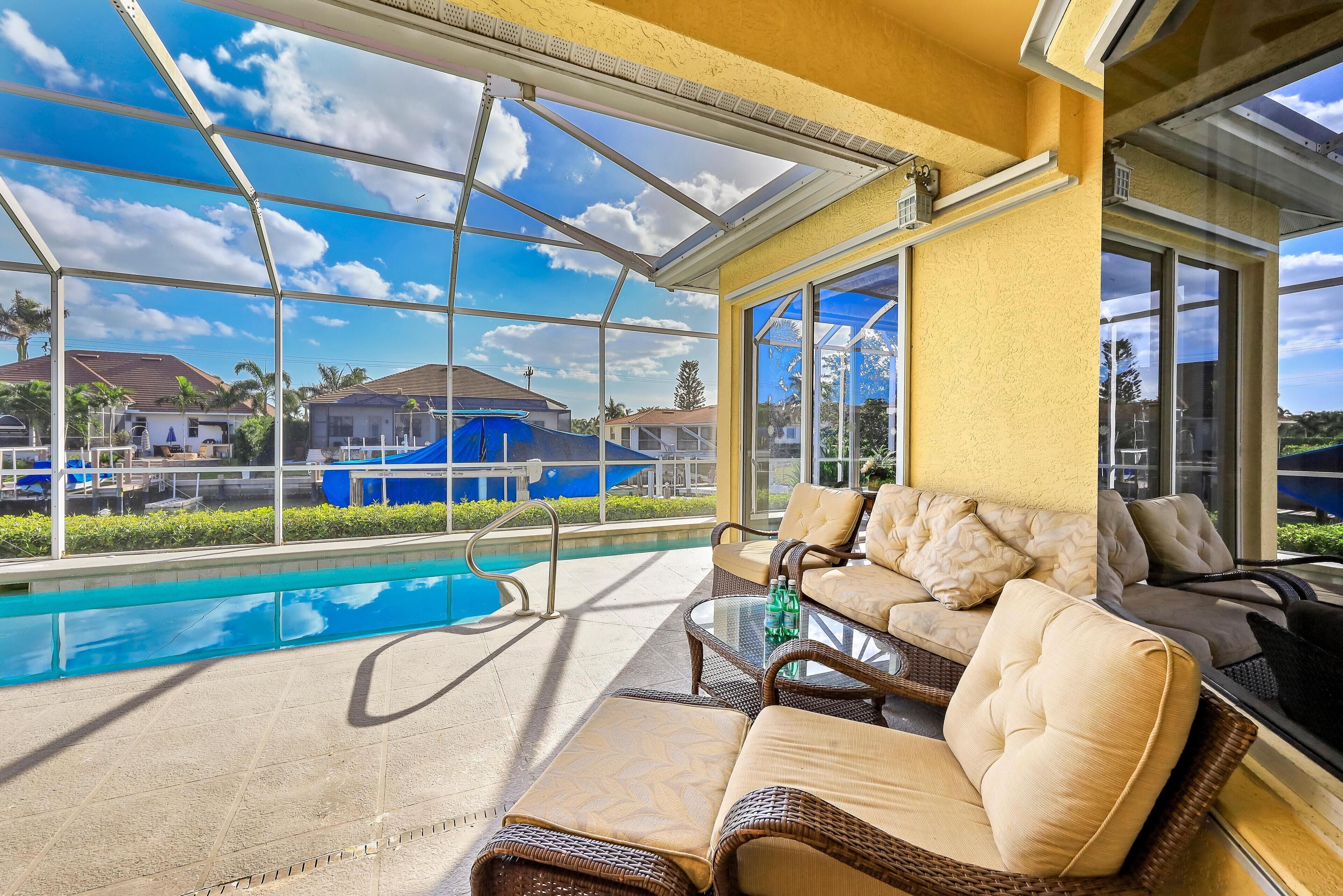 A sunlit outdoor area with a pool, wicker furniture, and large windows offering a view of the neighborhood.