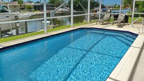 Una piscina con agua azul cristalina, rodeada por una terraza techada con sillas de descanso y vistas a barcos y casas al fondo.