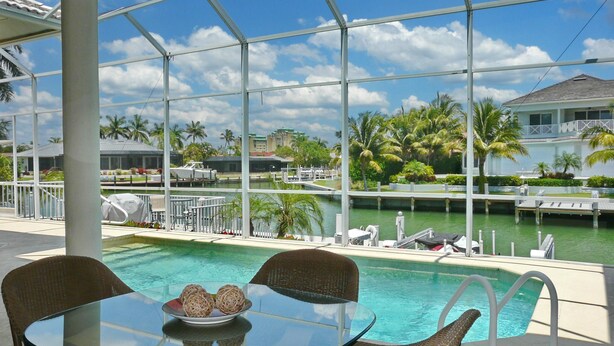 A glass-top table with wicker chairs and a decorative bowl, overlooking a swimming pool with a view of a marina and palm trees. - 931 Hyacinth Court (Marco Island)