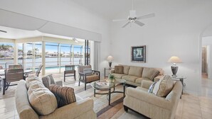 A spacious living room with a large window overlooking a pool and palm trees, a ceiling fan, and a comfortable seating area with a coffee table. - Unique Waterfront Home With Pool, spa and Guesthouse (Marco Island)