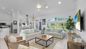 A modern living room with a sofa, coffee table, and chairs, a kitchen in the background, and a large window with a view of greenery. - Luxuriously Furnished Waterfront Home With Pool/spa (Marco Island)