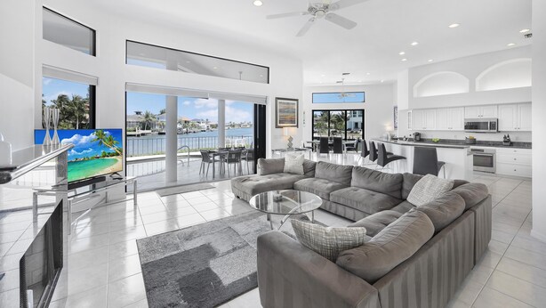 A spacious living area with a large sectional sofa, a glass coffee table, and a TV. There is a balcony with a view of palm trees and a building. - Waterfront Home on key lot With Pool, spa and Views (Marco Island)