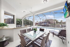 A sunlit patio with a glass-top table, wicker chairs, a pool, and a view of a residential area. - Elegantly Decorated Waterfront Home With Pool and spa (Marco Island)