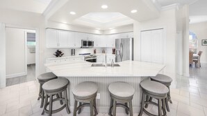 A modern kitchen with white cabinetry, a large island with bar stools, stainless steel appliances, and recessed ceiling lighting.