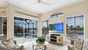 A spacious living room with a large TV, comfortable seating, a coffee table, and a view of a pool and outdoor area.