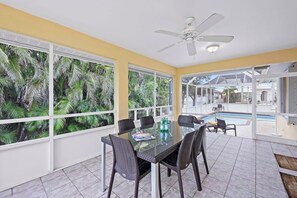 A dining area with a glass table and chairs, large windows with a view of palm trees, and a ceiling fan. - Pet-friendly Waterfront With Pool and Steps to Beach (Marco Island)