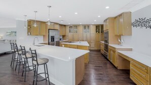 A modern kitchen with a central island, bar stools, wooden cabinets, and a white countertop. - 216 Shadowridge Court (Marco Island)
