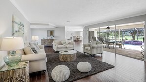 A spacious living room with a large window, a dining area, and a kitchen in the background. - 216 Shadowridge Court (Marco Island)