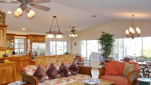 A spacious living room with a sofa, coffee table, and dining area. There is a kitchen with wooden cabinets and a refrigerator. The room has ceiling fans and pendant lights. - 848 Buttonwood Court (Marco Island)