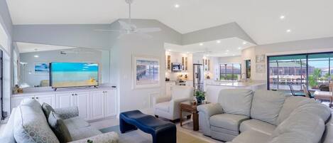 A spacious living area with a large sofa, a coffee table, and a TV. There is a kitchen in the background with white cabinets and a dining area with a table and chairs.