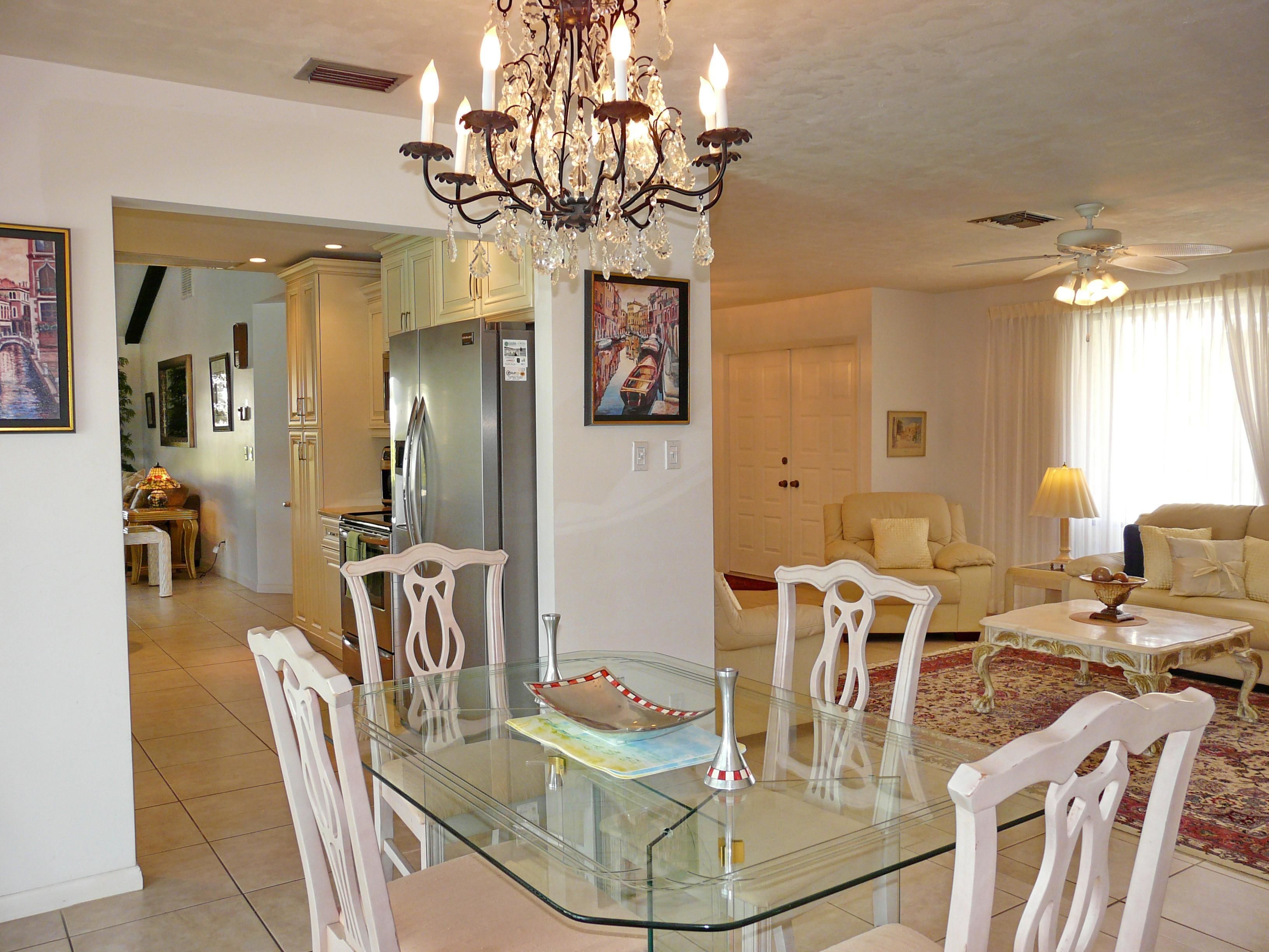 A dining area with a glass table, white chairs, a chandelier, and a kitchen in the background.