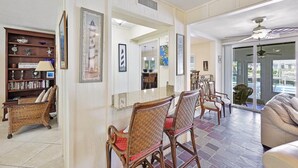 A spacious living area with a dining table, wicker chairs, a bookshelf, and a large window offering a view of the outdoors. - 775 Willow Court (Marco Island)