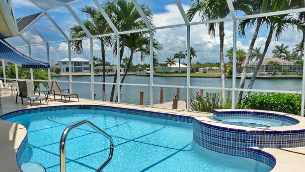 An indoor swimming pool with clear blue water, surrounded by a white metal frame and a view of palm trees and a body of water. - 1684 Rainbow Court (Marco Island)