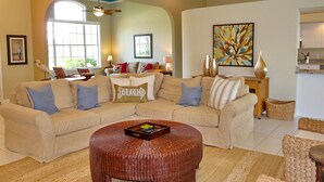 A living room with a beige sectional sofa, a round wicker coffee table, and a large window providing natural light. - 1684 Rainbow Court (Marco Island)