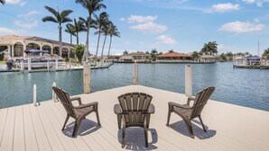 A deck with three Adirondack chairs overlooking a marina with boats and palm trees.
