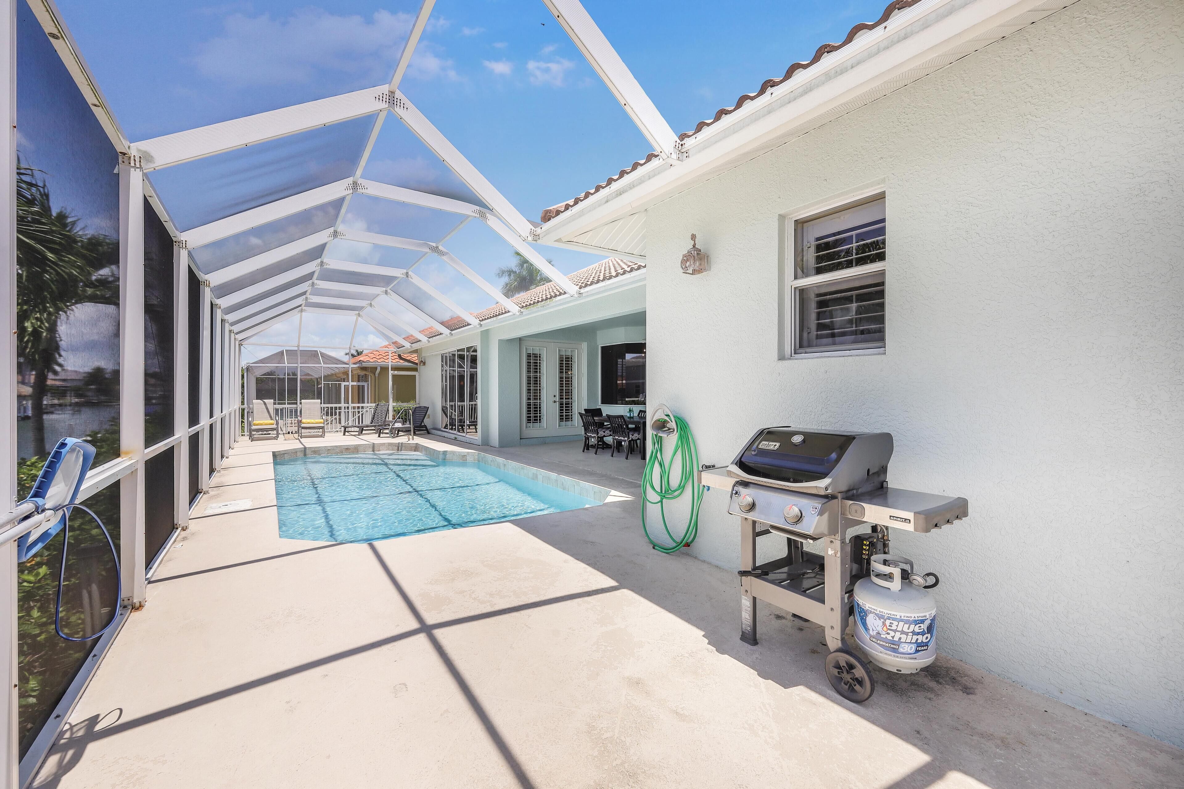 A covered outdoor swimming pool area with a barbecue grill and propane tank, surrounded by a white building with windows and a view of a body of water.
