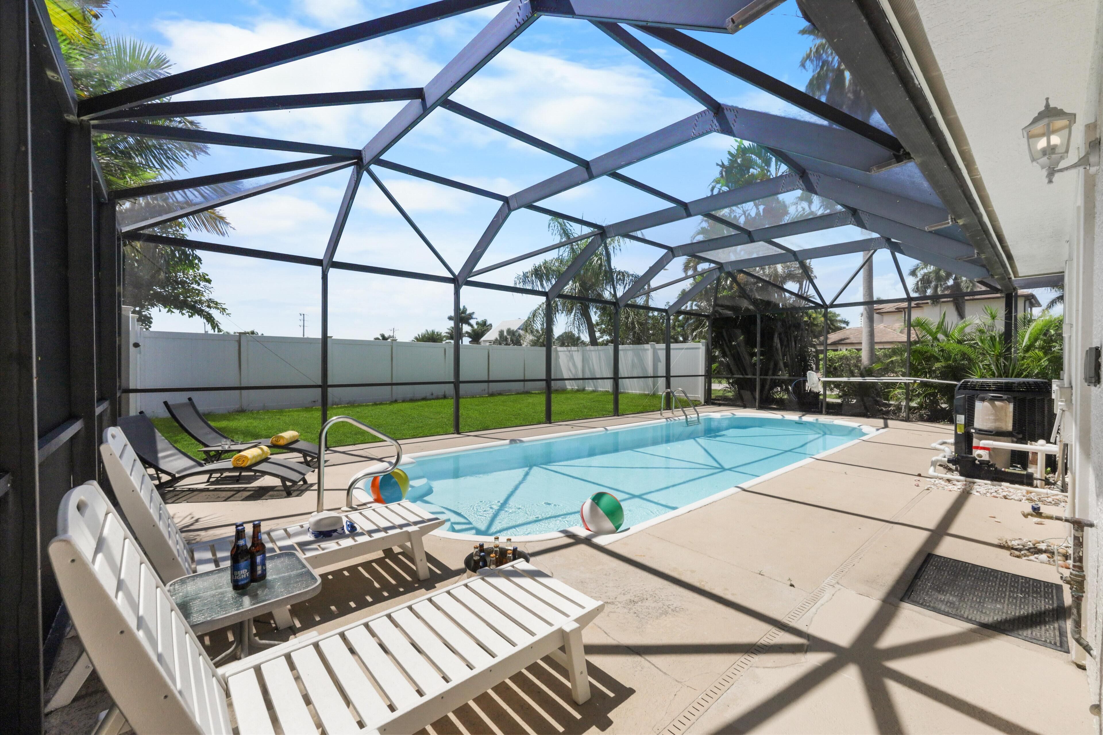 A covered outdoor swimming pool area with lounge chairs and a table, surrounded by a metal fence and palm trees.