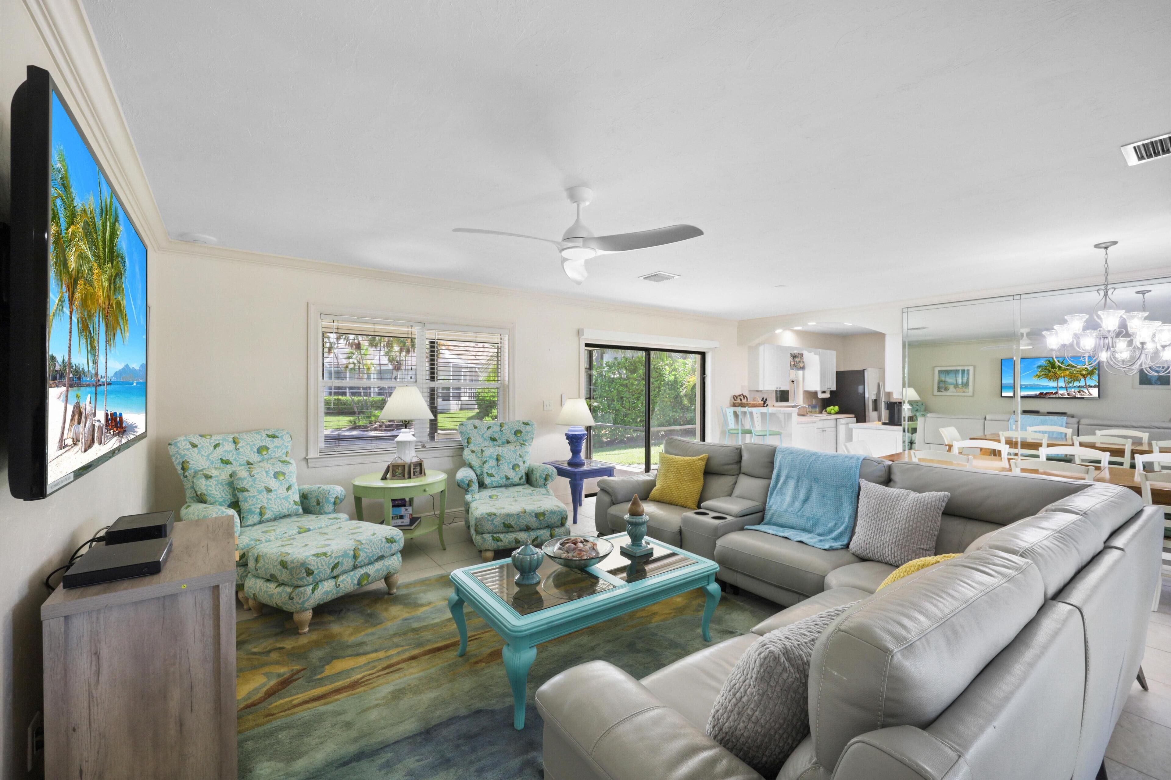 A spacious living room with a grey sofa, teal armchairs, and a coffee table. There is a ceiling fan, a large window with blinds, and a view of the outdoors.