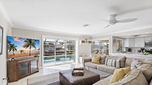 A spacious living room with a large TV, a leather ottoman, and a sofa with cushions. There is a kitchen area with a microwave and a dining table. - Waterfront Home With Pool and Western Exposure (Marco Island)